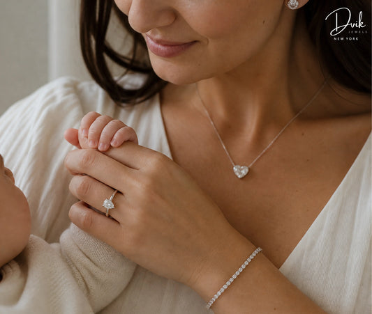 Dvik Jewels featuring a silver heart shaped diamond necklace, pear-cut diamond ring, and a tennis bracelet worn by a mother holding her baby's hand