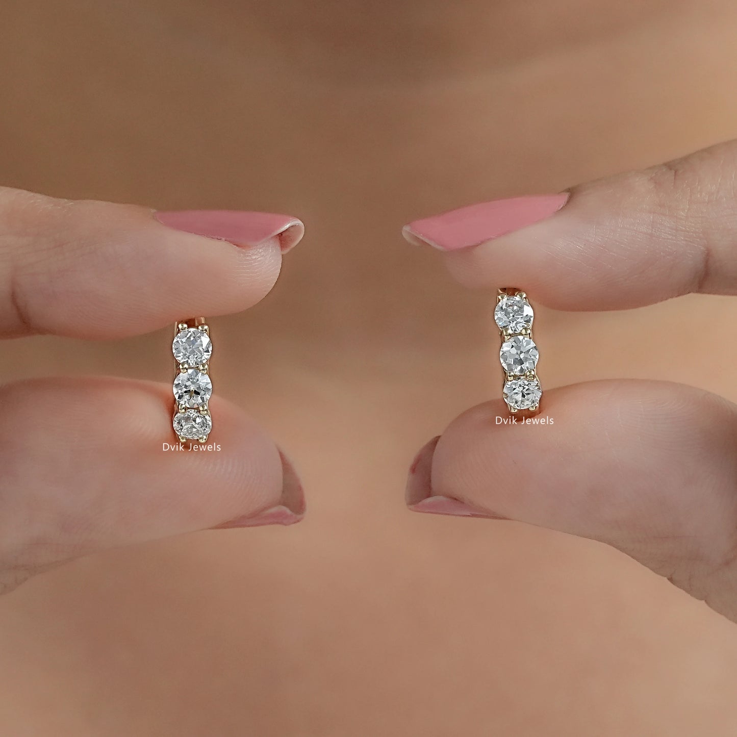 Pair of three stone huggie hoops held vertically between fingertips to showcase brilliant round-cut stones