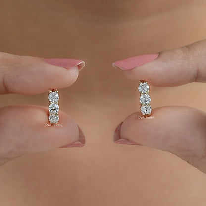 Pair of three stone huggie hoops held vertically between fingertips to showcase brilliant round-cut stones